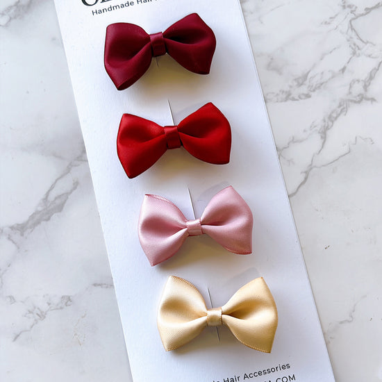 Four satin hair bows in burgundy, bright red, light pink, and pale yellow, displayed on a white Olive & Shea branded card over a light marble surface.