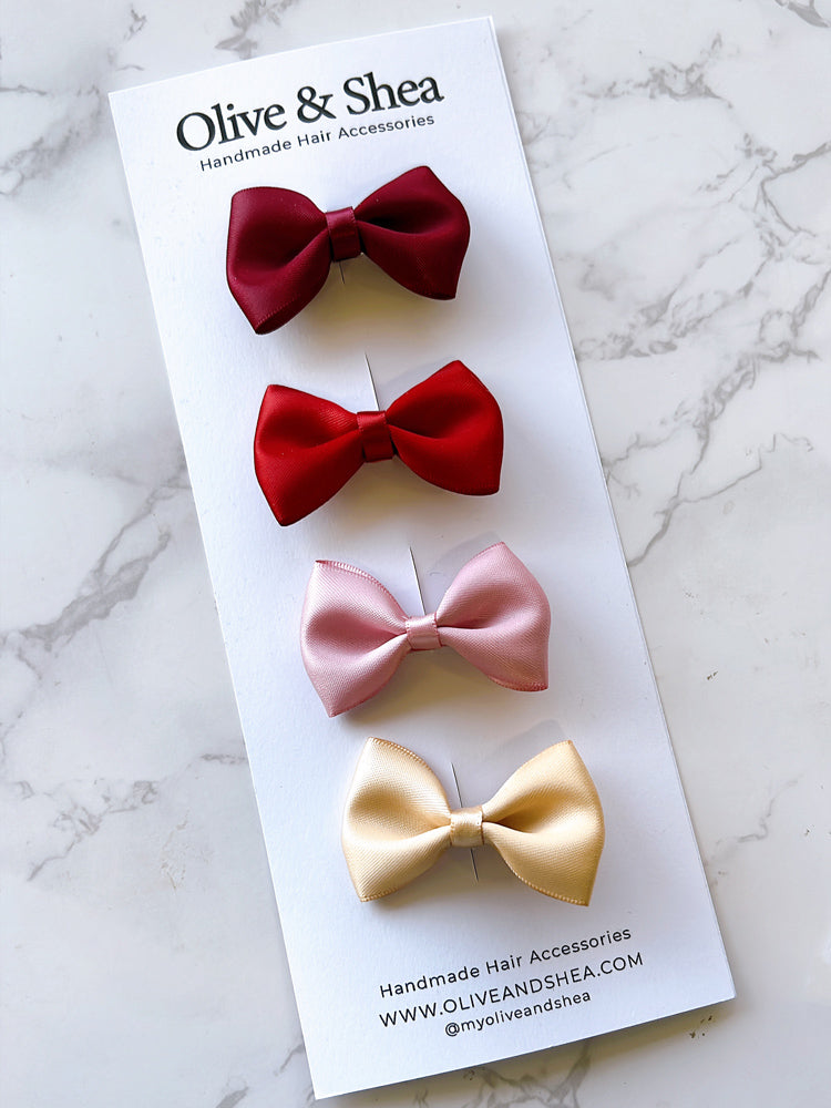 Four satin hair bows in burgundy, bright red, light pink, and pale yellow, displayed on a white Olive & Shea branded card over a light marble surface.