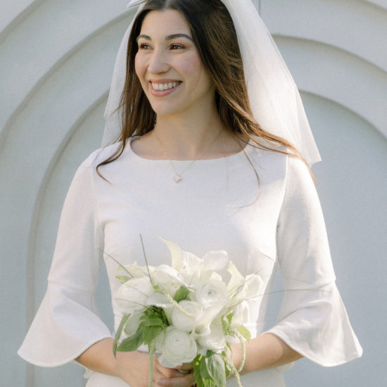 Woman in a white wedding dress with a veil and bouquet, smiling outdoors.