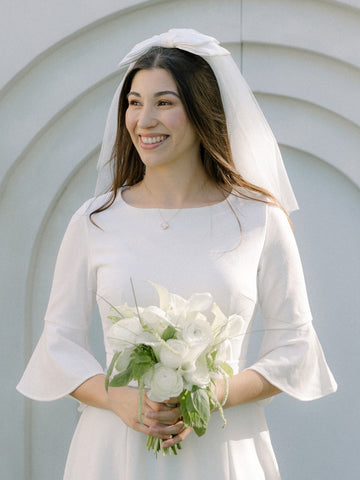 Woman in a white wedding dress with a veil and bouquet, smiling outdoors.