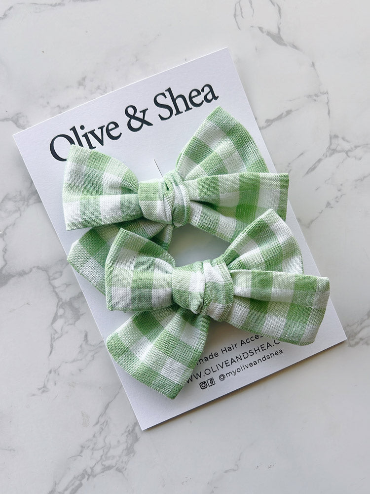Green gingham hair bows on a white card with 'Olive & Shea' branding on a marble background.