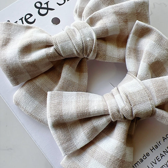 Two beige fabric hair bows on a white background with branding text.