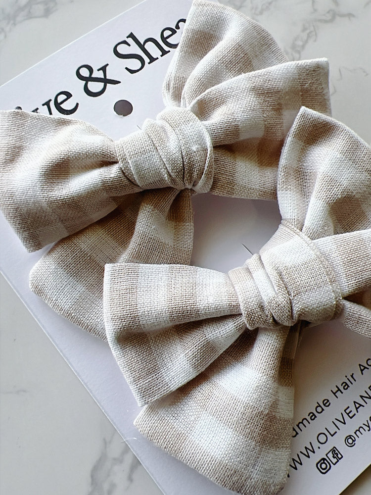 Two beige fabric hair bows on a white background with branding text.