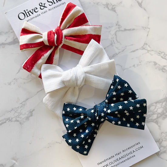 Three hair bows in red, white, and blue on a marble surface with Olive & Shea branding.
