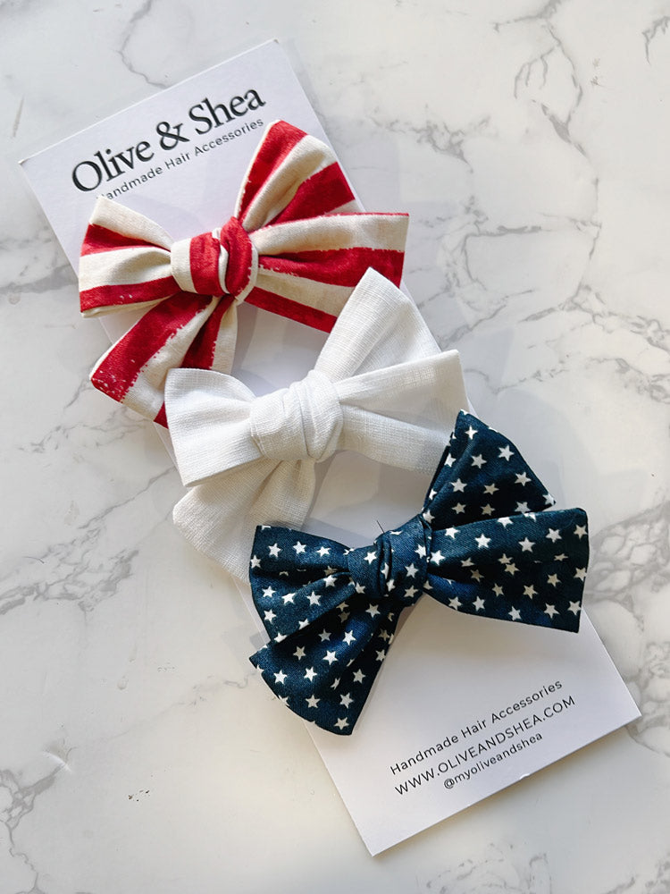 Three hair bows in red, white, and blue on a marble surface with Olive & Shea branding.