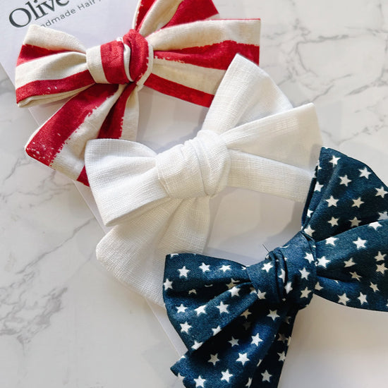 Three fabric hair bows in red, white, and blue on a marble surface with 'Olive & Shea' branding.