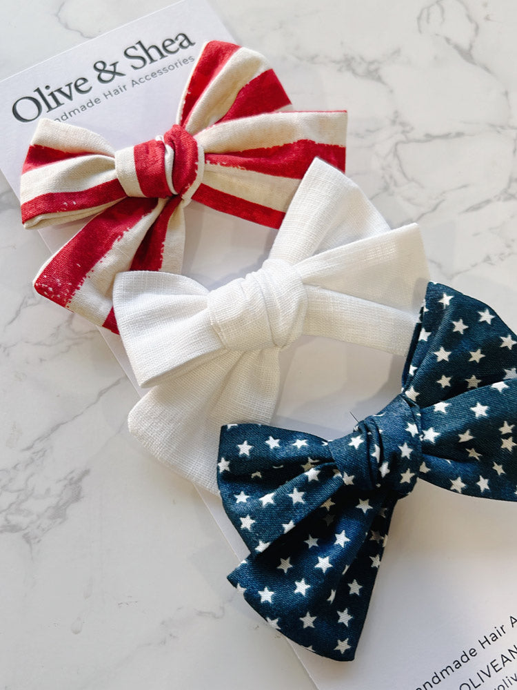 Three fabric hair bows in red, white, and blue on a marble surface with 'Olive & Shea' branding.